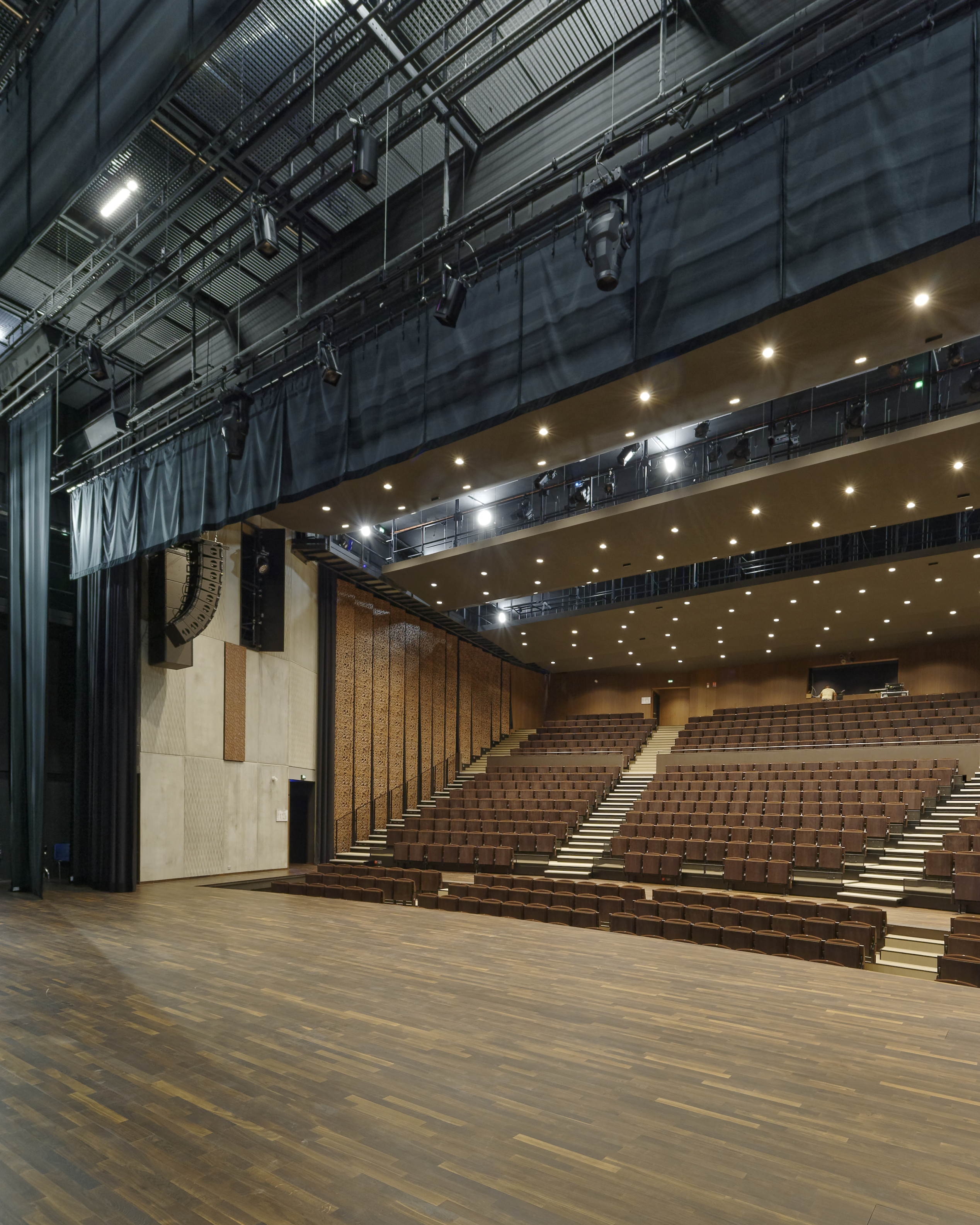 Salle de spectacle vue depuis le plateau : parquet, gradins et balcon, projecteurs au plafond
