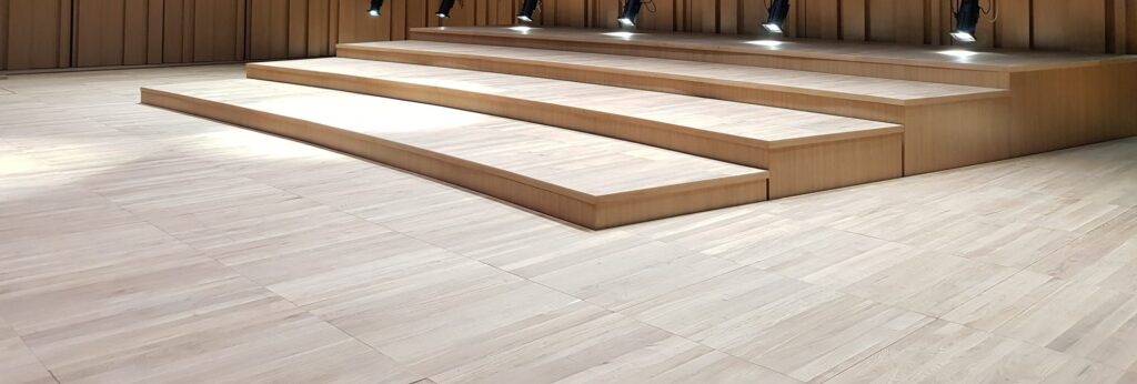 Gradins en bois et parquet de chêne sur une scène habillée de panneaux de bois, sous des projecteurs